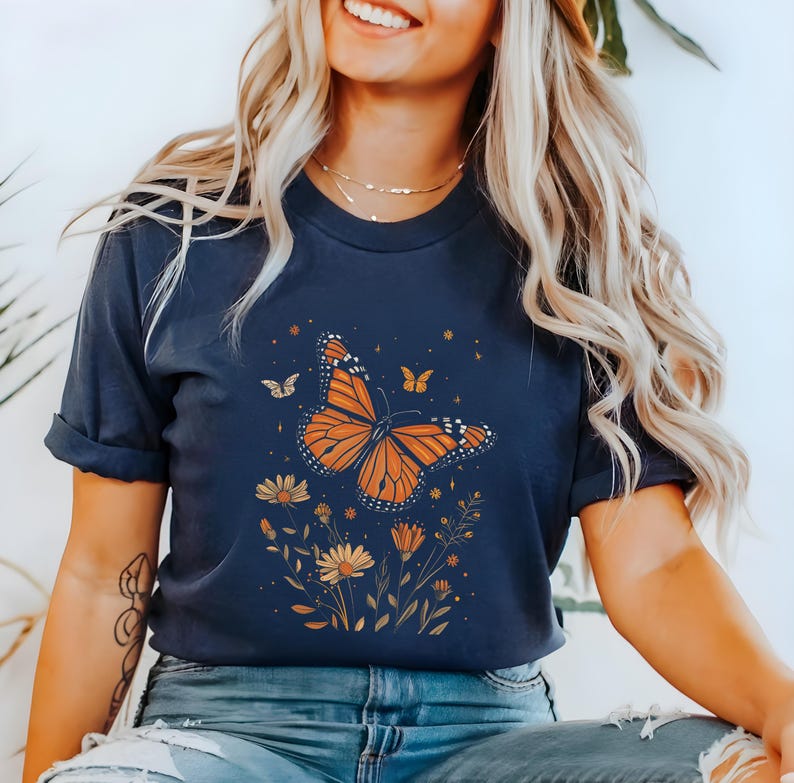 K&ouml;nnte beinhalten: Ein marineblaues T-Shirt mit einem Aufdruck eines orange-schwarzen Schmetterlings, umgeben von gelben Blumen und gr&uuml;nen Bl&auml;ttern. Der Schmetterling hat ein detailliertes Design und ist vor einem dunkelblauen Hintergrund platziert.