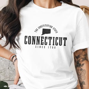 Puede incluir: Camiseta blanca con un diseño gráfico negro que presenta el estado de Connecticut, el texto "CONNECTICUT" y "SINCE 1788". La camiseta también incluye el texto "THE CONSTITUTION STATE" en un arco sobre el contorno del estado.
