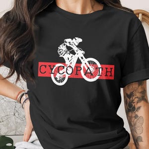 Può includere: T-shirt nera con grafica bianca di un ciclista su mountain bike. La parola "CYCOPATH" è stampata in bianco su un rettangolo rosso. La maglietta è realizzata in materiale morbido.