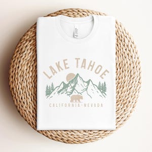 Puede incluir: Camiseta blanca con un dise&ntilde;o gr&aacute;fico de una cordillera con un oso y el texto "Lake Tahoe California - Nevada".