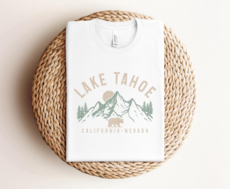 Puede incluir: Camiseta blanca con un dise&ntilde;o gr&aacute;fico de una cordillera con un oso y el texto "Lake Tahoe California - Nevada".