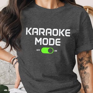 May include: Dark gray t-shirt with the words "KARAOKE MODE" in white block letters. Below the text is a green slider graphic with "OFF" and "ON" labels. The shirt is a casual style.