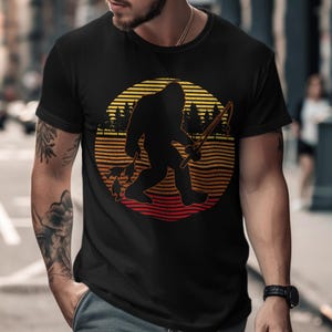 Pode incluir: Camiseta preta com uma silhueta de Bigfoot segurando uma vara de pescar e peixes, em um design de pôr do sol com listras laranja, amarelas e vermelhas. O design inclui uma silhueta de floresta.