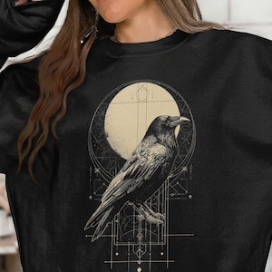 May include: A black crewneck sweatshirt featuring a graphic print of a raven perched in front of a large, pale circular moon. The design includes intricate geometric lines and celestial patterns in a light beige color, centered on the chest.