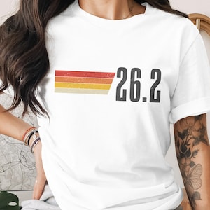 May include: White t-shirt with a retro design featuring a horizontal stripe in red, orange, yellow, and beige, with the number "26.2" in bold, black font. The shirt is likely for marathon runners.