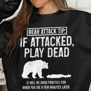 May include: Black sweatshirt with white text: "BEAR ATTACK TIP: IF ATTACKED, PLAY DEAD." A white bear illustration stands over a person lying down. The text below reads: "IT WILL BE GOOD PRACTICE FOR WHEN YOU DIE A FEW MINUTES LATER."