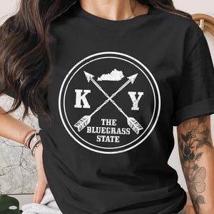 Może przedstawiać: Czarny t-shirt z białym okrągłym wzorem. Wzór zawiera skrzyżowane strzały, litery "K" i "Y" oraz tekst "The Bluegrass State". Zawiera również sylwetkę stanu Kentucky.