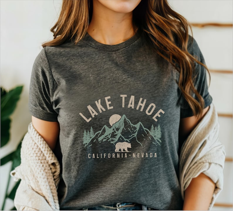 Puede incluir: Una camiseta gris oscuro con un gr&aacute;fico de estilo vintage de monta&ntilde;as, un oso y un sol. El texto "LAKE TAHOE CALIFORNIA-NEVADA" est&aacute; impreso en la camiseta.