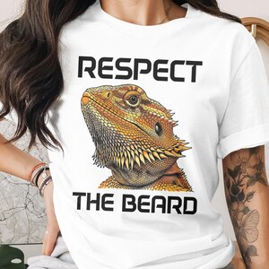 May include: White t-shirt featuring a detailed illustration of a bearded dragon in shades of orange, yellow, and brown. The text "RESPECT THE BEARD" is printed above and below the reptile image in bold, black capital letters.
