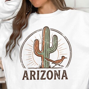 May include: A white crewneck sweatshirt featuring a graphic print of a saguaro cactus and a roadrunner bird inside a circular frame with sunburst lines. The word ARIZONA is printed in large brown capital letters below the circular design.