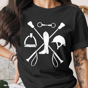 May include: Black t-shirt featuring a white equestrian design. The graphic includes crossed riding crops, a stirrup, a riding boot, a helmet, and a horse bit. This design is perfect for horse riding enthusiasts.