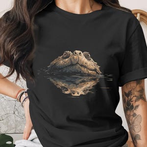 May include: A black short-sleeved t-shirt featuring a graphic print of a snapping turtle head emerging from the water. The turtle is depicted in shades of brown and tan, with a clear reflection visible on the water surface below the animal's chin.