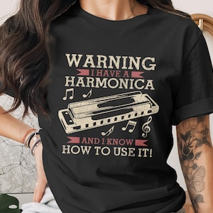 May include: Black t-shirt with the text "WARNING I HAVE A HARMONICA AND I KNOW HOW TO USE IT!" in a vintage style. The design includes a harmonica and musical notes in a beige color.