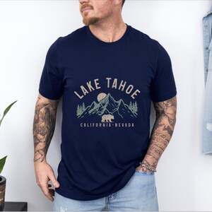 Puede incluir: Una camiseta azul marino con un dise&ntilde;o gr&aacute;fico de monta&ntilde;as, un oso y una luna. El texto "LAKE TAHOE CALIFORNIA-NEVADA" est&aacute; impreso en la camiseta.