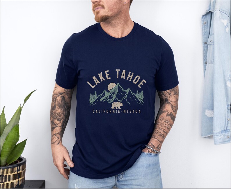 Puede incluir: Una camiseta azul marino con un dise&ntilde;o gr&aacute;fico de monta&ntilde;as, un oso y una luna. El texto "LAKE TAHOE CALIFORNIA-NEVADA" est&aacute; impreso en la camiseta.