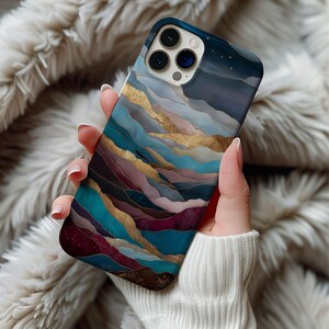 May include: A smartphone case featuring a landscape design with layers of teal, gold, purple, and brown. The case has a dark blue top with small white stars. The phone case is held in a person's hand.