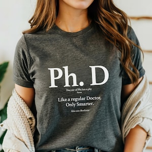 May include: Dark gray t-shirt with the text "Ph. D" in large white letters. Below the text is the definition of Ph.D. and the phrase "Like a regular Doctor, Only Smarter." The shirt is a crew neck.
