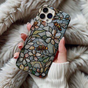 May include: A smartphone case featuring a stained-glass design with bees and flowers. The case has a light blue background with floral and bee motifs in shades of white, yellow, and brown. The case is held in a person's hand.