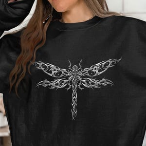 May include: A black long-sleeve sweatshirt featuring a large, intricate, metallic silver dragonfly graphic centered on the chest. The dragonfly design is composed of sharp, flowing tribal-style lines and curves that create a reflective, chrome-like appearance.