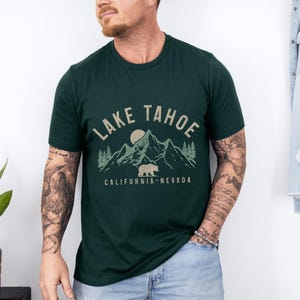 Puede incluir: Una camiseta verde oscuro con un dise&ntilde;o gr&aacute;fico de monta&ntilde;as, un oso y una luna. El texto "Lake Tahoe" est&aacute; escrito en marr&oacute;n por encima del gr&aacute;fico. El texto "California - Nevada" est&aacute; escrito en marr&oacute;n por debajo del gr&aacute;fico.