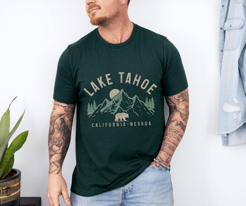 Puede incluir: Una camiseta verde oscuro con un dise&ntilde;o gr&aacute;fico de monta&ntilde;as, un oso y una luna. El texto "Lake Tahoe" est&aacute; escrito en marr&oacute;n por encima del gr&aacute;fico. El texto "California - Nevada" est&aacute; escrito en marr&oacute;n por debajo del gr&aacute;fico.