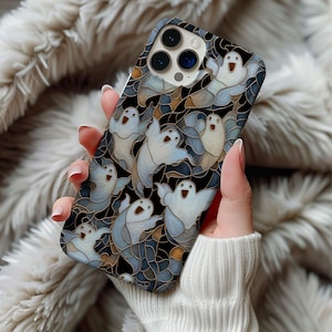 Puede incluir: Una funda de teléfono inteligente con un patrón repetido de fantasmas de dibujos animados en blanco, azul y gris sobre un fondo negro. El diseño tiene un efecto de vidriera. La funda está en la mano de una persona.