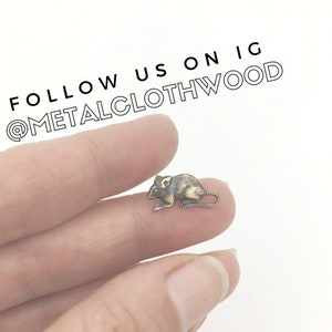 May include: A small, bronze-colored mouse figurine is held between two fingers. The figurine is detailed with black accents and has a curled tail. The text "FOLLOW US ON IG @METALCLOTHWOOD" is visible at the top of the image.