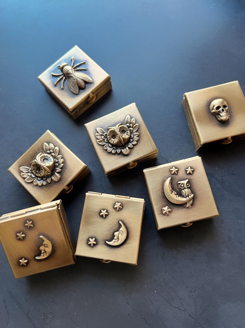 May include: Six small, square, brass-colored boxes with different designs. The designs include an owl, a crescent moon with stars, a skull, and a bee. The boxes are hinged and have a small latch.