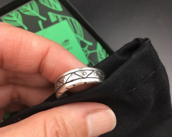 May include: A silver ring with a geometric pattern. The ring is on a finger and is being held over a black fabric pouch.