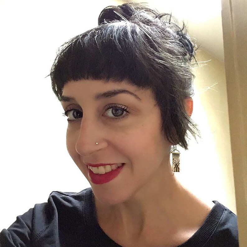 May include: A woman with short black hair and bangs smiles at the camera. She is wearing a black shirt and has a small silver nose ring. She is wearing a silver earring with a small, intricate design.