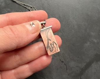 Pode incluir: Um colar de prata com um pingente que apresenta uma gravação detalhada de uma mantis religiosa em uma placa retangular de cobre.