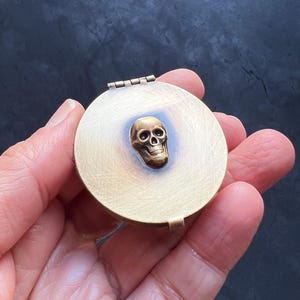 Könnte beinhalten: Eine runde Schatulle aus Messing mit einem Scharnierdeckel. Die Schatulle hat ein erhabenes, goldfarbenes Totenkopfdesign auf der Vorderseite.