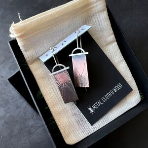May include: A pair of silver earrings with a black and pink centipede design. The earrings are packaged in a black box with a white cloth bag and a card that reads "Metal Cloth & Wood".
