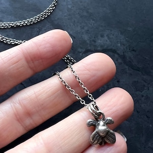 May include: A small, silver-toned bee pendant necklace. The bee charm has detailed wings and body. The chain is a delicate, linked design. The necklace is displayed against a dark background.