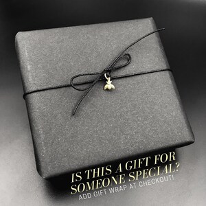 May include: A black gift box with a black ribbon tied around it. A small gold bee charm hangs from the ribbon. The box has the text "IS THIS A GIFT FOR SOMEONE SPECIAL? ADD GIFT WRAP AT CHECKOUT!" printed on it.