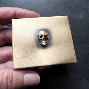 May include: A small, square, brass box with a raised, three-dimensional skull design. The box has a brushed finish and is closed.
