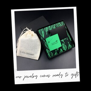 May include: A black gift box with a green leaf pattern and a green card that says "Caring for You MCAW Jewelry". The box is sitting on a white surface. The box is also accompanied by a small white drawstring bag with the words "METAL, CLOTH & WOOD" printed on it.