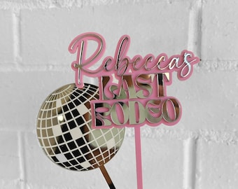 May include: Pink acrylic cake topper with the text "Rebecca's Last Fling" in gold lettering and a silver disco ball.