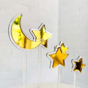 May include: A set of gold acrylic cake toppers featuring a crescent moon and three stars. The moon and stars are translucent and have a mirrored gold finish.