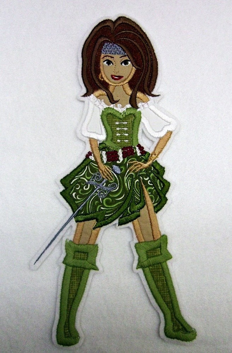 K&ouml;nnte beinhalten: Gesticktes Abzeichen einer Piratin in einem gr&uuml;n-wei&szlig;en Outfit, die ein Schwert h&auml;lt. Die Piratin hat braunes Haar, ein blaues Stirnband und goldene Creolen. Das Design weist komplizierte Details auf.