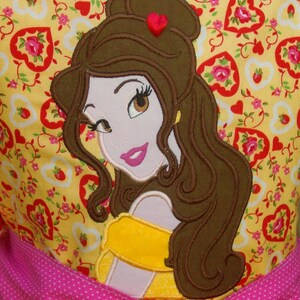 May include: Embroidered applique of Belle from Beauty and the Beast on a yellow fabric with a pink and white polka dot ribbon. The fabric is covered in a pattern of red hearts and flowers.