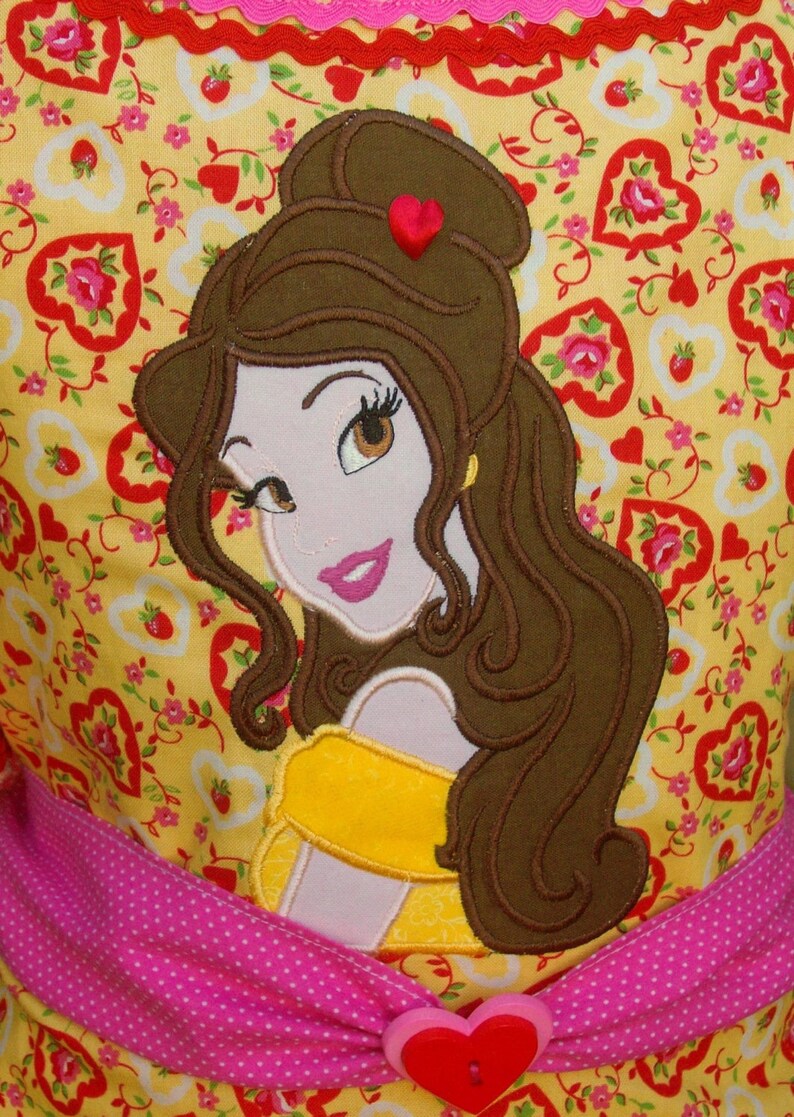 May include: Embroidered applique of Belle from Beauty and the Beast on a yellow fabric with a pink and white polka dot ribbon. The fabric is covered in a pattern of red hearts and flowers.