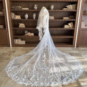Puede incluir: Un velo de novia blanco largo con intrincados detalles de encaje floral. El velo se exhibe en un maniquí, mostrando su longitud y delicado diseño. El ribete de encaje presenta un patrón repetido de hojas y flores.