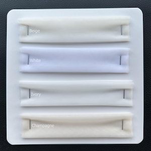May include: A white card showing fabric swatches in Beige, White, Ivory, and Champagne. Each fabric strip is pinned with a silver pin. The text labels each colour. The background is dark.