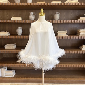 May include: A white cape with a square neckline and a feathery trim. The cape is displayed on a mannequin with a gold-coloured neck. The background features a wooden bookshelf with books and decorative vases.