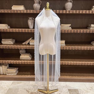 以下が含まれることがあります： 繊細なパール装飾が施された、透け感のある白いウェディングベール。 金色のスタンドにマネキンが飾られており、その流れるようなデザインが際立っています。 ベールの縁には小さなパールが飾られ、エレガントさを添えています。