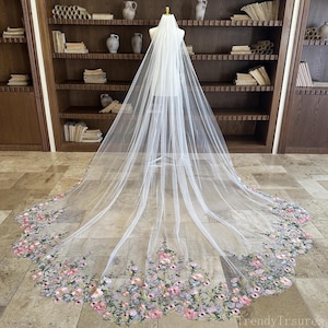 May include: A long, white bridal veil with a floral embroidered border. The veil is displayed on a mannequin and features a delicate, flowing design. The embroidered flowers are in shades of pink, purple, and green.