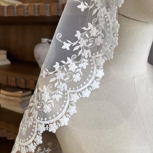 May include: A white bridal veil with a floral lace trim. The lace is adorned with small flowers and leaves, forming a scalloped edge. The veil is draped over a mannequin, highlighting the intricate design and texture of the fabric.