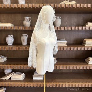Op de afbeelding: Een cr&egrave;mekleurige kanten sluier, versierd met gedetailleerde bloempatronen en een geschulpte rand, wordt getoond op een mannequin. De mannequin staat tegen een achtergrond van houten planken gevuld met boeken en aardewerk.