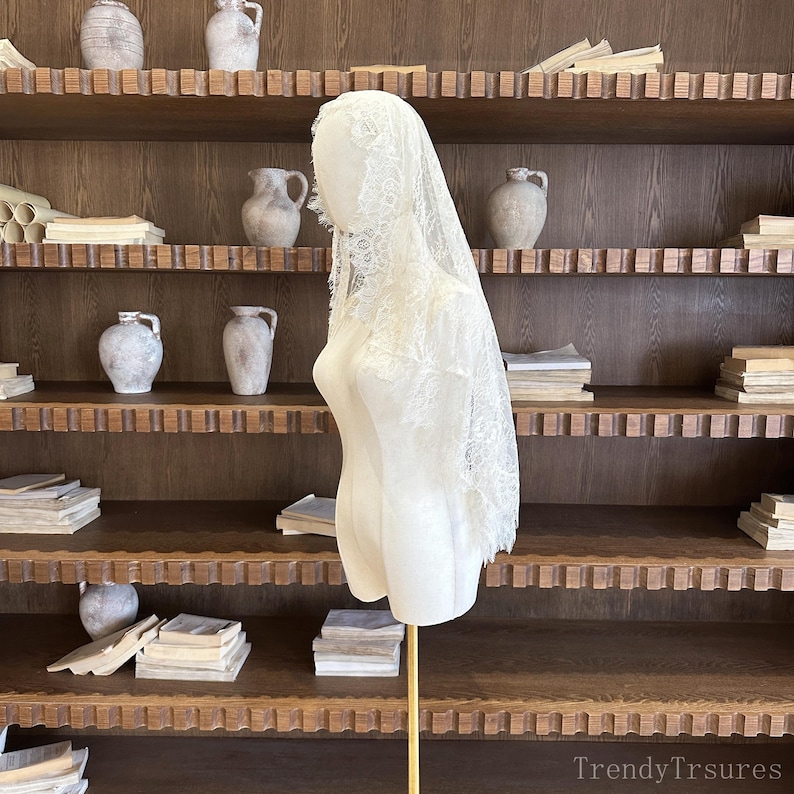 Op de afbeelding: Een witte kanten sluier op een mannequin. De sluier, met zijn gedetailleerde bloemenpatroon, is over het hoofd en de schouders van de mannequin gedrapeerd. De mannequin staat op een gouden standaard, geplaatst tegen een houten boekenplank met diverse decoratieve items.
